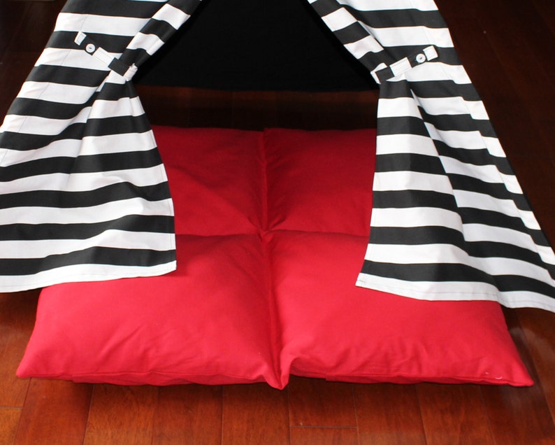 May include: A child's play tent with a black and white striped canopy and a bright red floor cushion. The tent has a teepee shape and is set on a wooden floor.