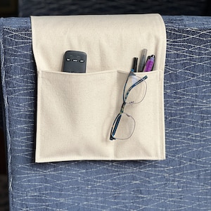 May include: A beige fabric armrest organizer with two pockets. The organizer has a remote control, glasses, and pens in the pockets.