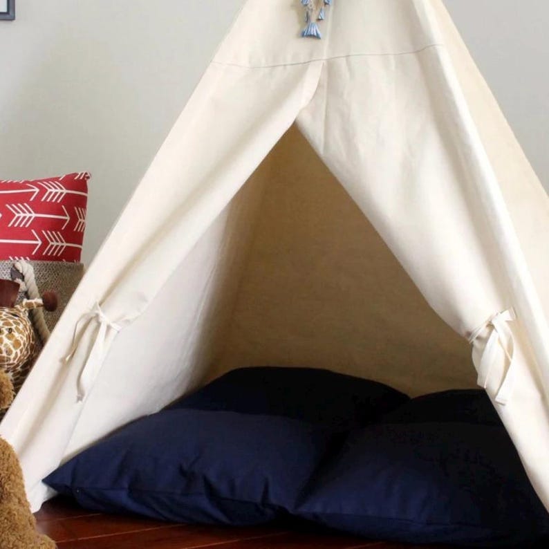 May include: A cream-colored teepee tent with a navy blue cushion inside. The tent has a simple design with ties and a decorative element at the top. A red pillow with white arrows is visible in the background.