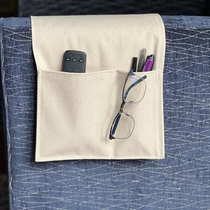 May include: A cream-colored fabric armrest organizer featuring two pockets draped over a blue textured surface. The left pocket holds a black remote control, while the right pocket contains a pair of eyeglasses and three pens.
