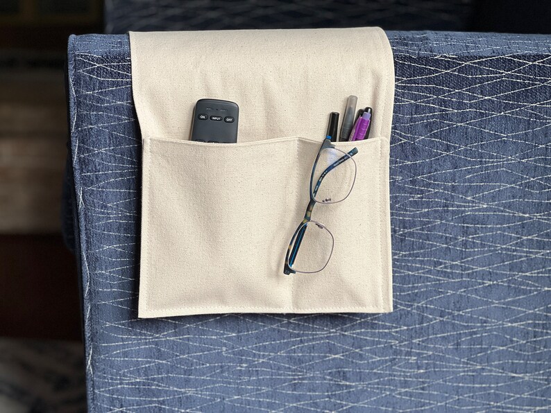 May include: A cream-colored fabric armrest organizer featuring two pockets draped over a blue textured surface. The left pocket holds a black remote control, while the right pocket contains a pair of eyeglasses and three pens.