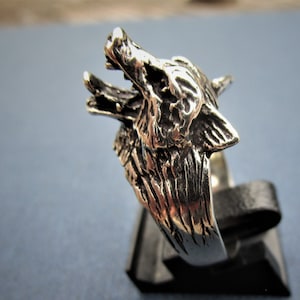 Puede incluir: Anillo plateado con la cabeza de un lobo detallada, con la boca abierta. La joya se muestra sobre un fondo oscuro, revelando los detalles del pelaje y los rasgos del lobo. Una pieza de joyería.
