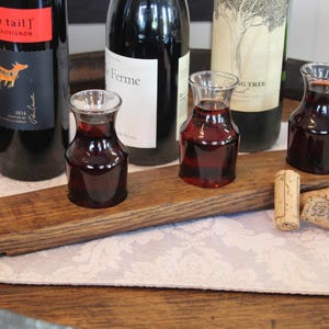 May include: A wooden wine tasting tray with three small glass carafes filled with red wine. The tray is sitting on a white tablecloth with a floral pattern. There are three wine bottles in the background, two with labels that read "Yellow Tail" and "Ferme".