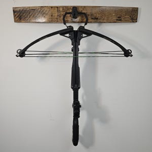 May include: A black crossbow hanging on a wooden plank. The crossbow has a black frame, limbs, and stock. The bowstring is black and green. The wooden plank is mounted on a white wall.