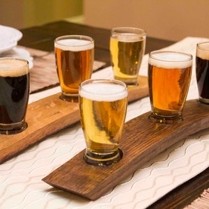 May include: A wooden beer flight with five glasses filled with different types of beer. The glasses are arranged in a row on the flight, which is made of dark wood. The flight is sitting on a white table with a white tablecloth.