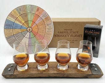 May include: A whiskey flight set featuring four clear Glencairn glasses filled with amber liquid, resting on a curved wooden barrel stave base with metal accents. Behind the glasses is a circular flavor wheel chart and a cardboard box.