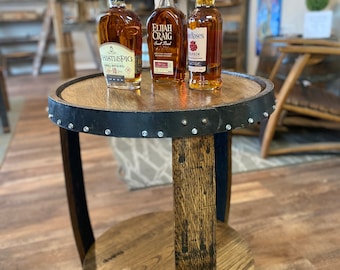 May include: A wooden barrel table with a round top and a lower shelf. Three bottles of whiskey are displayed on the top shelf. The bottles are labeled "WhistlePig", "Elijah Craig", and "Roses".