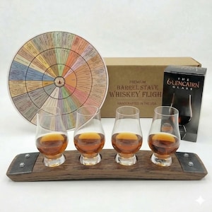 May include: A whiskey tasting set featuring four Glencairn glasses filled with amber liquid, resting on a wooden barrel stave. A flavor wheel and a box labeled "Premium Barrel Stave Whiskey Flight" are also present. The set includes a box of "The Glencairn Glass".