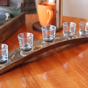 May include: A wooden shot glass holder with a curved design. The holder is made from reclaimed wood and has a dark brown stain. Five clear shot glasses are placed in the holder.