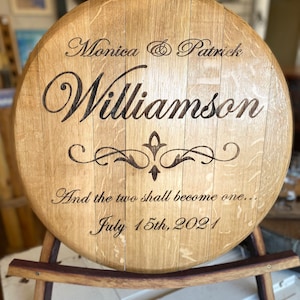 May include: A round wooden sign with the names "Monica & Patrick Williamson" and the date "July 15th, 2021" engraved on it. The sign also includes the phrase "And the two shall become one..." and is displayed on a wooden stand.