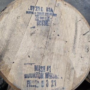 May include: A circular wooden barrel head with natural light brown grain. Black stenciled text reads LOT 21E 03A, BUFFALO TRACE DISTILLERY, MASH #1, BOURBON WHISKEY, and FILLED 5 3 21. The surface shows visible wood planks joined together with dark markings.