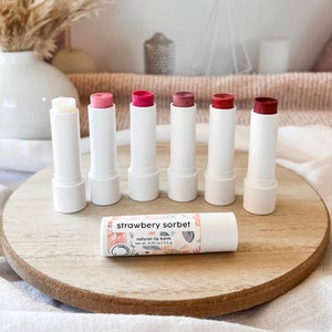 May include: Six white tubes of natural lip balm with different shades of pink and red on a wooden surface. The lip balm tubes are labeled "strawberry sorbet" and "natural lip balm".