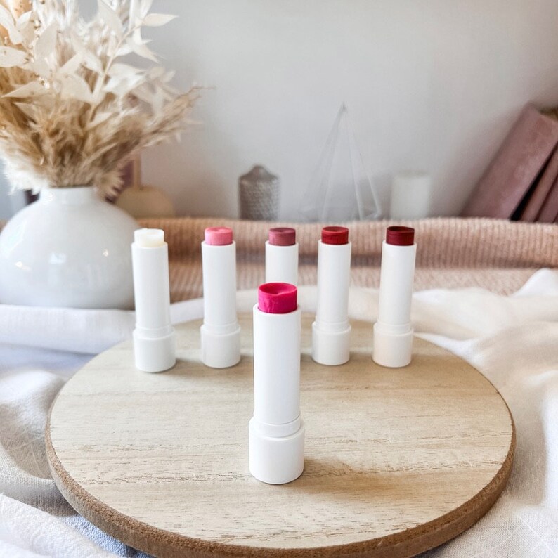 May include: A set of seven lip balms in various shades of pink and red, arranged on a wooden surface. The lip balms are in white tubes with a clear cap.