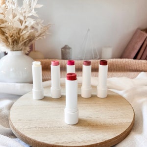 May include: A set of seven lip balms in white tubes with different colored tops. The lip balms are arranged in a circle on a wooden surface.