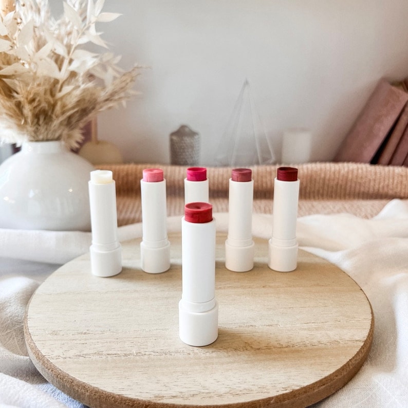 May include: A set of seven lip balms in white tubes with different colored tops. The lip balms are arranged in a circle on a wooden surface.