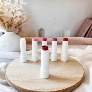 May include: A set of six lip balms in various shades of pink and red, arranged on a wooden tray. The lip balms are in white tubes with a white cap.