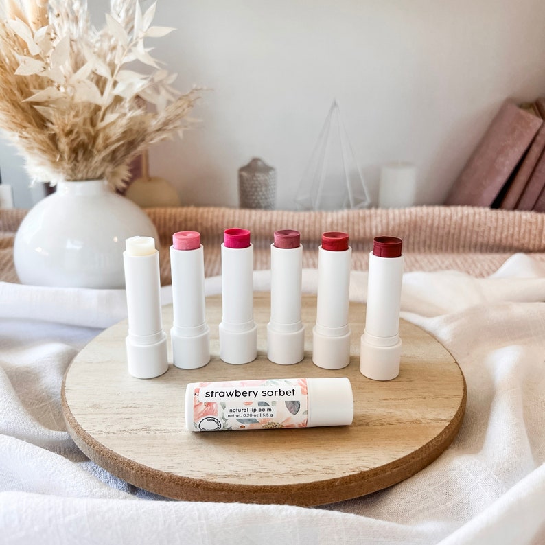 May include: Six white tubes of natural lip balm with different shades of pink and red on a wooden surface. The lip balm tubes are labeled "strawberry sorbet" and "natural lip balm".