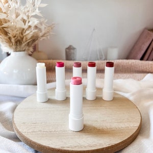 May include: Six white tubes of lip balm with different colored tints. The tubes are arranged in a semi-circle on a wooden surface.