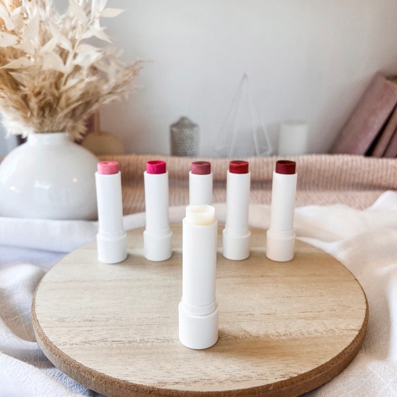 May include: Six white lip balm tubes with different colored tops. The tubes are arranged in a circle on a wooden surface. One tube is open and shows a white balm inside.