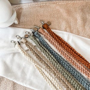 Puede incluir: Una colección de cordones trenzados en varios colores, incluyendo crema, beige, verde salvia, gris azulado, melocotón y óxido. Cada cordón tiene un cierre plateado para sujetar llaves o una tarjeta de identificación. Los cordones están colocados sobre una tela blanca.