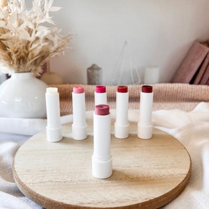 May include: Six lip balms in white tubes with different colored tops. The tubes are arranged in a row on a wooden surface. The colors of the tops are: clear, pink, red, pink, red, and red.