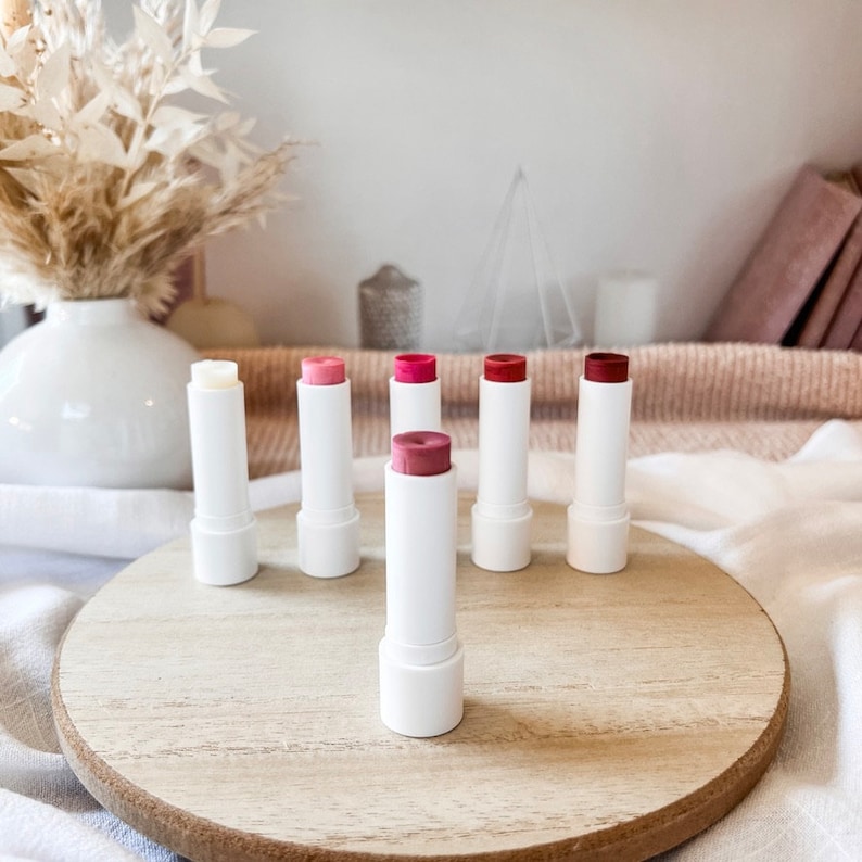 May include: Six lip balms in white tubes with different colored tops. The tubes are arranged in a row on a wooden surface. The colors of the tops are: clear, pink, red, pink, red, and red.