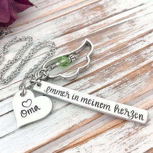May include: Silver necklace with a heart charm that says "Oma" and a bar charm that says "immer in meinem herzen". The necklace also has a small silver wing charm with a green crystal.