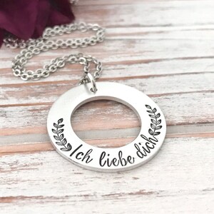 May include: Silver necklace with a round pendant that says "Ich liebe dich" in black lettering. The pendant has a small circle cut out of the center and is surrounded by a wreath of leaves.