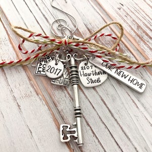 May include: A silver key ornament with a house charm, a "Sold" sign charm, a "2017" charm, and a heart charm with the text "2107 Hawthorn Street". The ornament also has a tag that reads "Our New Home".