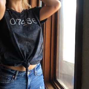 Puede incluir: Una camiseta gris oscuro con el número "07456" impreso en la parte delantera. La camiseta está anudada a la cintura y combinada con vaqueros azules. La modelo está de pie cerca de un marco de ventana de madera.