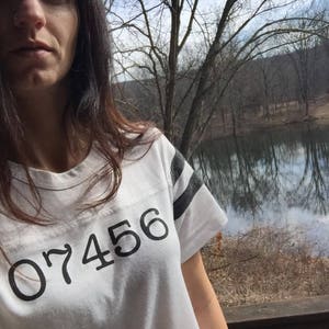 Puede incluir: Camiseta blanca con rayas negras en las mangas y el número "07456" impreso en la parte delantera. La camiseta se usa al aire libre con un fondo natural de árboles y agua.