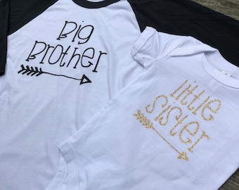 Puede incluir: Una camiseta blanca y negra estilo béisbol con las palabras "Big Brother" en escritura negra y un gráfico de flecha. Al lado, un body blanco con "Little Sister" en escritura dorada con purpurina y un gráfico de flecha. Ropa para bebés y niños pequeños.