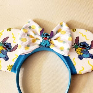 May include: A blue headband with white ears featuring a cartoon character holding a pineapple. The ears have a yellow and white pineapple pattern. A matching bow and character embellishment complete the design.