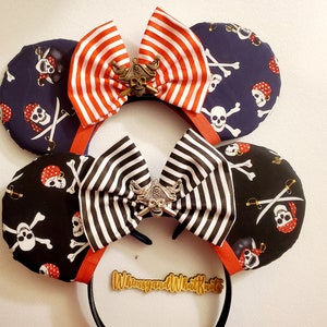 May include: A pair of black and white striped fabric Minnie Mouse ears with a gold skull and crossbones charm. The ears are decorated with a black and white striped bow and a red and white striped bow. The ears are on a black headband.