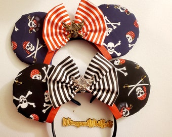 May include: A pair of black and white striped fabric Minnie Mouse ears with a gold skull and crossbones charm. The ears are decorated with a black and white striped bow and a red and white striped bow. The ears are on a black headband.