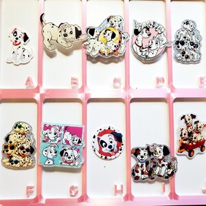 May include: A collection of ten acrylic pins featuring various Dalmatian puppy illustrations, arranged in a pink grid organizer labeled with letters A through J. Each pin displays different poses, including puppies in a wagon, wearing sunglasses, or playing.