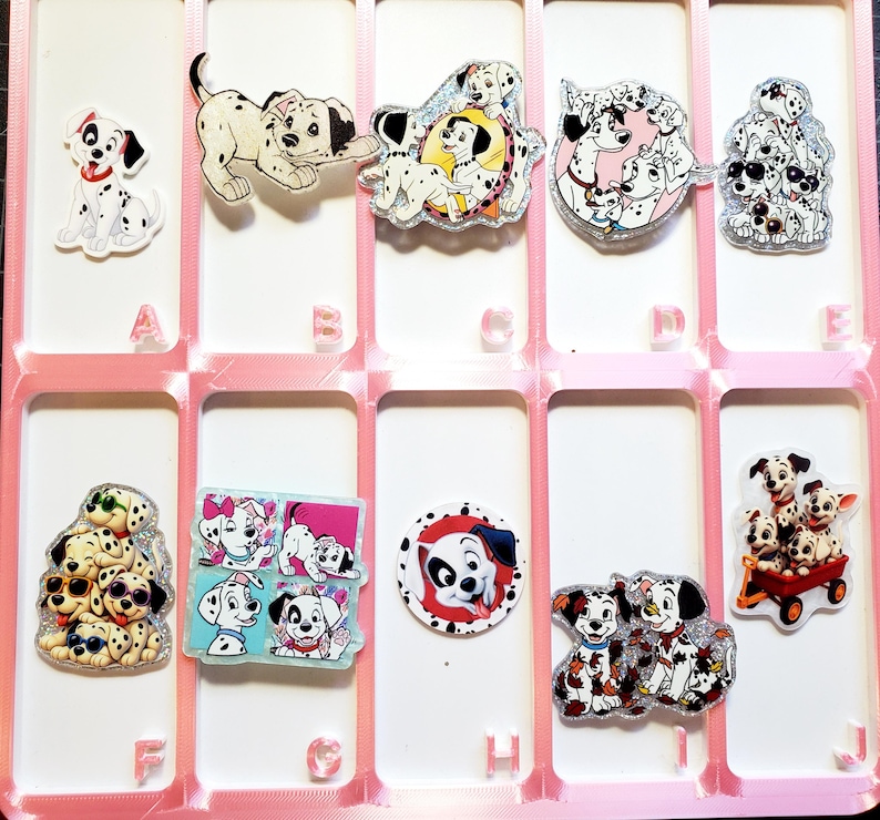 May include: A collection of ten acrylic pins featuring various Dalmatian puppy illustrations, arranged in a pink grid organizer labeled with letters A through J. Each pin displays different poses, including puppies in a wagon, wearing sunglasses, or playing.