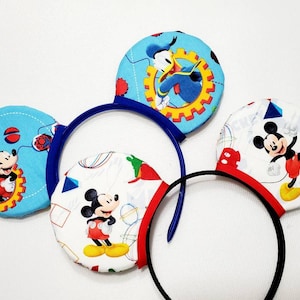 May include: A pair of blue and black Mickey Mouse ears with a blue, white and red fabric design featuring Mickey Mouse and Donald Duck. The ears are decorated with a geometric pattern and the text "Mickey Mouse Clubhouse".