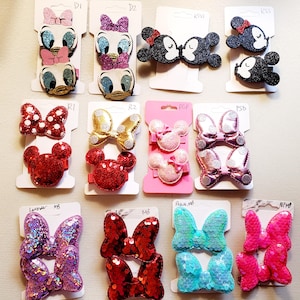 May include: A collection of colorful hair clips featuring Disney characters and bow designs. The clips include images of Mickey and Minnie Mouse, Daisy Duck, and bows in various colors like red, pink, and blue. The clips are attached to white cards.