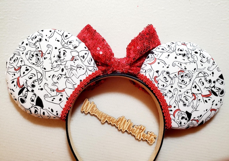May include: A headband featuring two circular ears covered in a white fabric with a black and white dalmatian puppy print. A bright red sequined bow sits between the ears, accented with red braided trim. A wooden cutout reads WhimsyandWhatKnots.