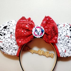 Può includere: Una fascia per capelli con grandi orecchie rotonde con un motivo dalmata in bianco e nero. Un grande fiocco rosso con paillettes è centrato sulla fascia, con un'immagine circolare di un cane dalmata. La fascia per capelli ha il testo "Whimsy and What Knots".