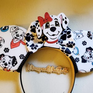 May include: A headband featuring two circular ears with a white fabric cover printed with black and white dalmatian puppies and paw prints. A central bow displays a dalmatian puppy face with a red glittery bow and collar. A wooden sign reads WhimsyandWhatKnots.