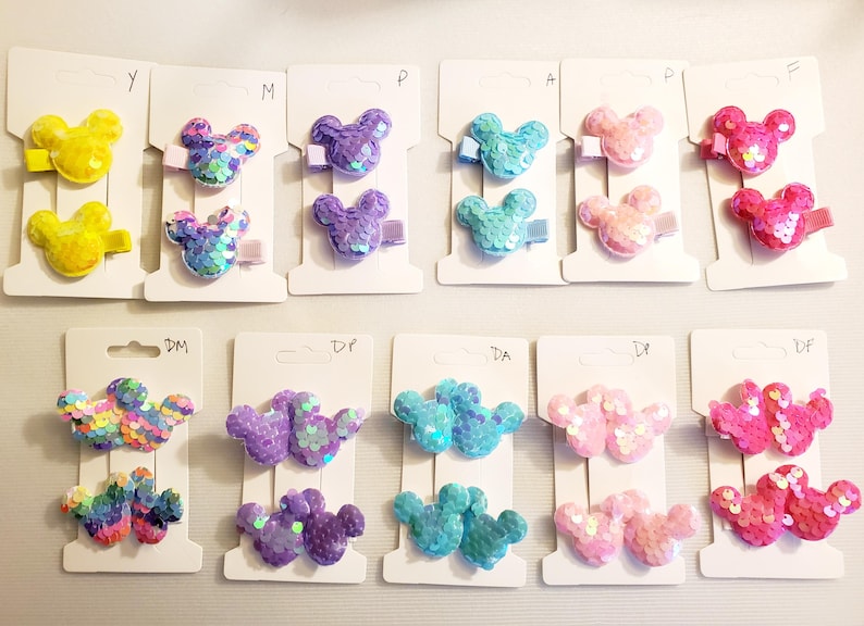 May include: Assortment of colorful Mickey Mouse hair clips with sequins. The clips are displayed on white cards with the letters Y, M, P, A, P, E, DM, DF, DA, DP, and DF.