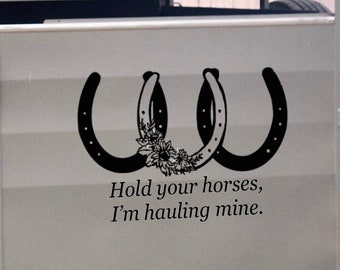 以下が含まれることがあります： 馬のトレーラーに、黒い馬蹄のステッカーが貼ってあります。馬蹄には花が描かれており、その下に「Hold your horses, I'm hauling mine.」という文字があります。トレーラーはグレーです。