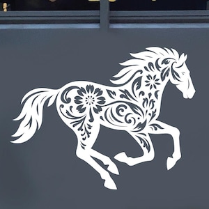 Könnte beinhalten: Ein weißer Pferdeaufkleber mit floralem Muster ist auf der Rückseite eines dunkelgrauen Pferdeanhängers angebracht. Das Design des galoppierenden Pferdes weist komplizierte Blumen- und Wirbeldetails auf, die einen dekorativen und künstlerischen Look erzeugen. Der Anhänger steht im Freien.