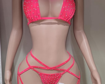 Puede incluir: Un conjunto de bikini rosa neón con pedrería. El conjunto incluye un top halter y una braguita de estilo tanga con tirantes a juego. El maniquí también lleva medias de rejilla rosas. La palabra "Rayen" está escrita en rosa neón.