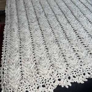 May include: A handmade, cream-colored crochet blanket with a chevron pattern. The blanket features a scalloped edge and is made with speckled yarn, adding visual interest. The blanket is likely for a bed or sofa.