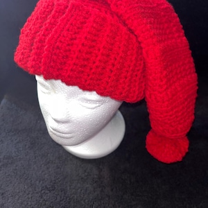 May include: A bright red, handmade crochet hat with a long, pointed tip and a small pom-pom at the end. The hat is displayed on a white mannequin head against a dark background. The hat's texture is clearly visible, showcasing the tight crochet stitches.