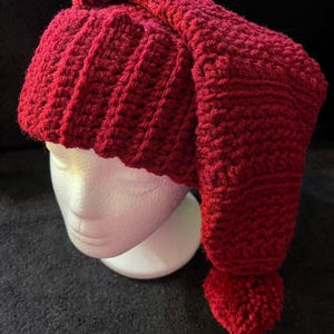 May include: A handmade, red crocheted hat with a long, pointed end and a small pom-pom. The hat is displayed on a white mannequin head against a dark background. The hat's texture is visible, showcasing the detailed crochet work.