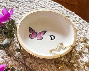 May include: A white ceramic trinket dish with a gold rim, featuring a pink butterfly and the letter "D". A gold chain necklace is resting on the dish.